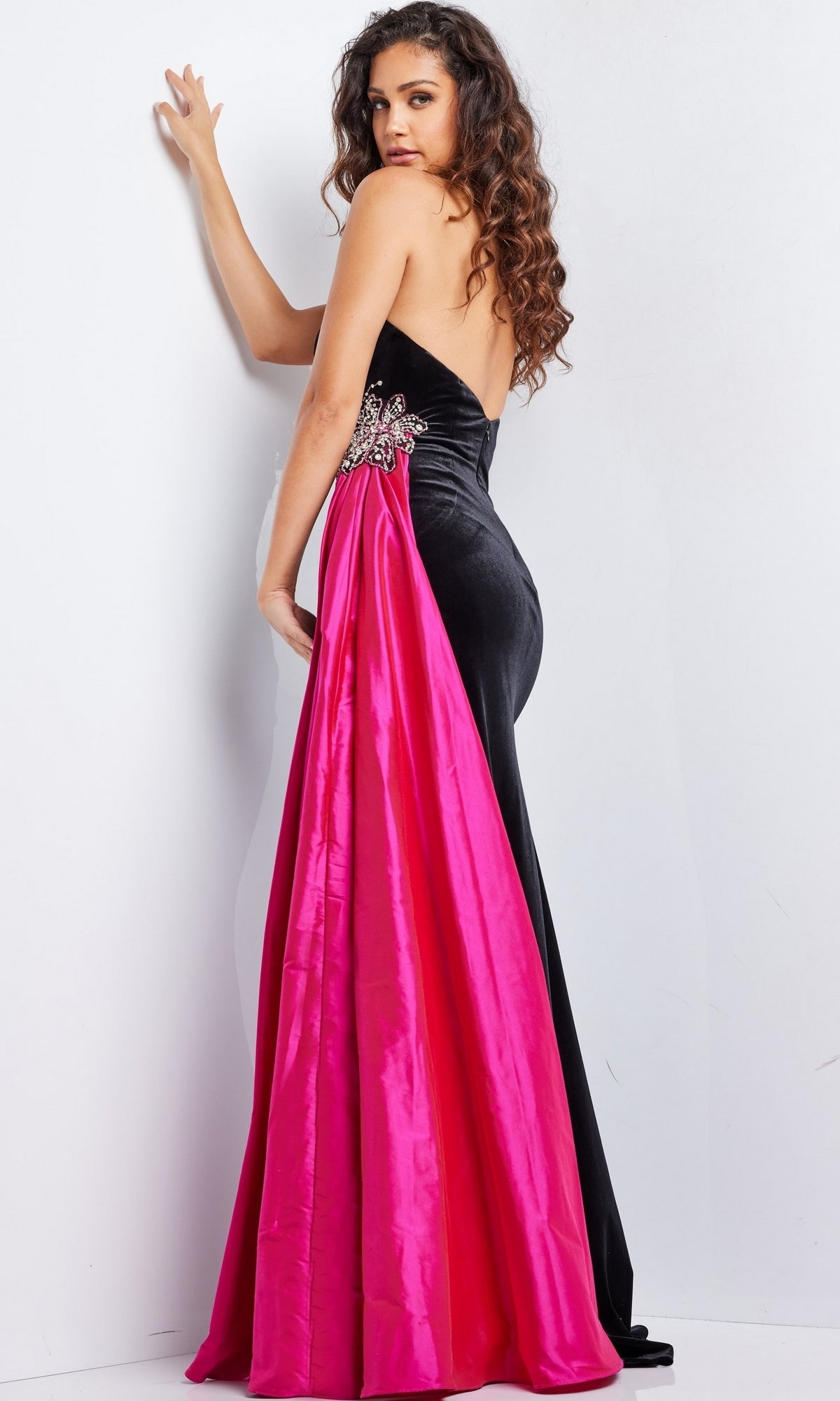 A woman models the Jovani Side-Sash Strapless Long Prom Dress 26117—featuring luxurious velvet, a bright pink satin train, and sparkling beadwork—while standing against a white wall and glancing over her shoulder.